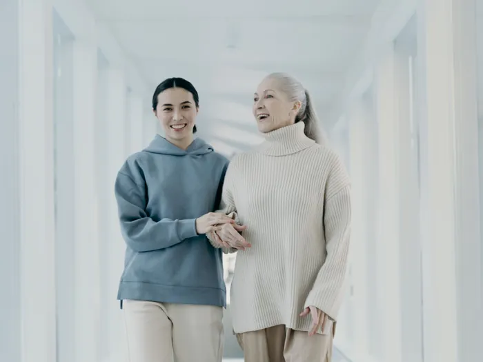 A caregiver and an older woman walking arm in arm down a bright hallway, smiling